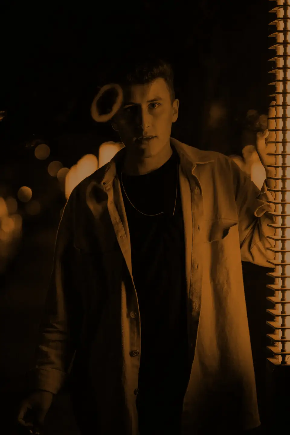 A man in an open overshirt and chain, city lights behind